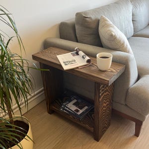 Puede incluir: Una mesa auxiliar de madera teñida oscura con un estante inferior. Una revista enrollada y una taza blanca están encima. La mesa está junto a un sofá gris y una planta en maceta. La mesa mide aproximadamente 61 cm de largo.