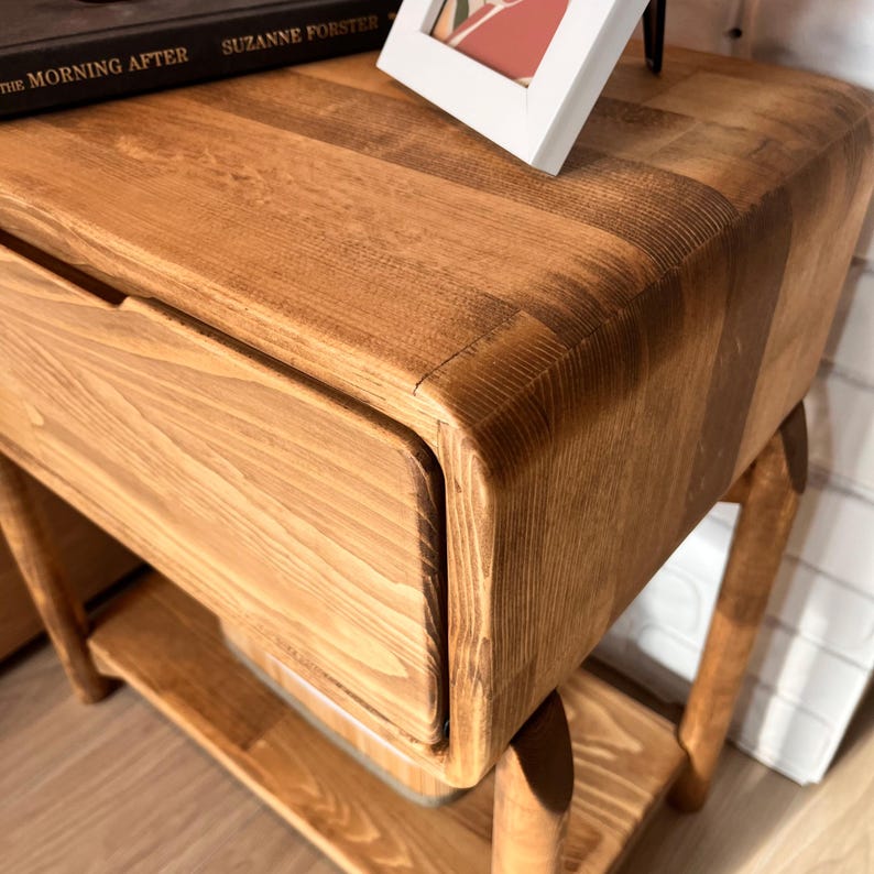 May include: A wooden nightstand with a drawer and a lower shelf. The nightstand is made of light brown wood and has rounded edges. A book and a framed picture are on top of the nightstand.