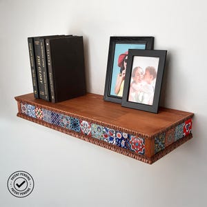 Könnte beinhalten: Ein schwebendes Holzregal mit einer dekorativen Bordüre aus bunten Fliesen. Das Regal enthält drei Bücher und zwei schwarz gerahmte Fotos. Das Regal ist an einer weißen Wand befestigt. Der Text "Patent Pending" ist sichtbar.