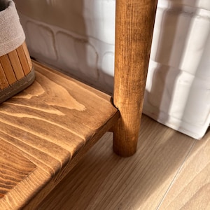 May include: Close-up of a wooden shelf with a warm, brown stain. The shelf has a natural wood grain pattern and is supported by a round, wooden leg. A woven basket is partially visible on the shelf.