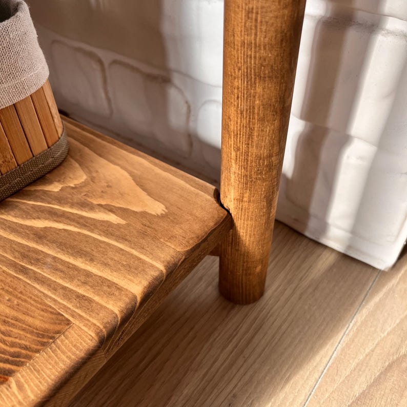May include: Close-up of a wooden shelf with a warm, brown stain. The shelf has a natural wood grain pattern and is supported by a round, wooden leg. A woven basket is partially visible on the shelf.