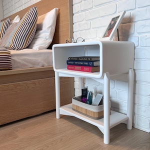 Puede incluir: Mesita de noche blanca con dos estantes. El estante superior tiene una pila de libros y el estante inferior tiene una cesta tejida con artículos de tocador. La mesa tiene bordes redondeados y un diseño moderno.