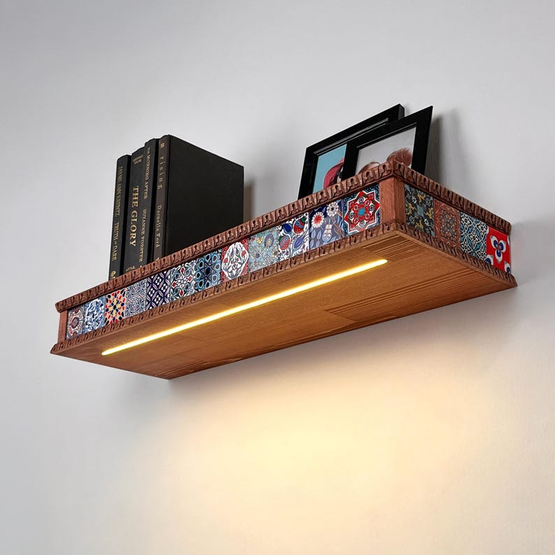 May include: A wooden wall shelf featuring a front edge decorated with a row of colorful patterned tiles and an integrated LED light strip. The shelf holds three black books with visible text and two black picture frames on its top surface.