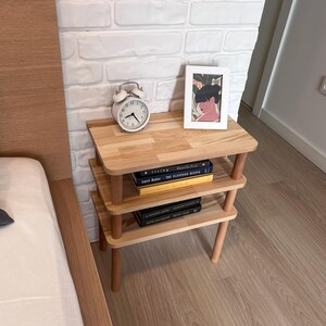 Puede incluir: Una mesita de noche de madera de tres niveles con esquinas y patas redondeadas. Un despertador blanco y una impresi&oacute;n enmarcada se encuentran en el estante superior. Los libros est&aacute;n apilados en los estantes inferiores. La mesita es de madera clara.