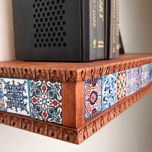 May include: A wooden floating shelf featuring a carved edge with repeating arch patterns and a front panel inlaid with square ceramic tiles displaying intricate blue, red, and white floral and geometric patterns, supporting black books.