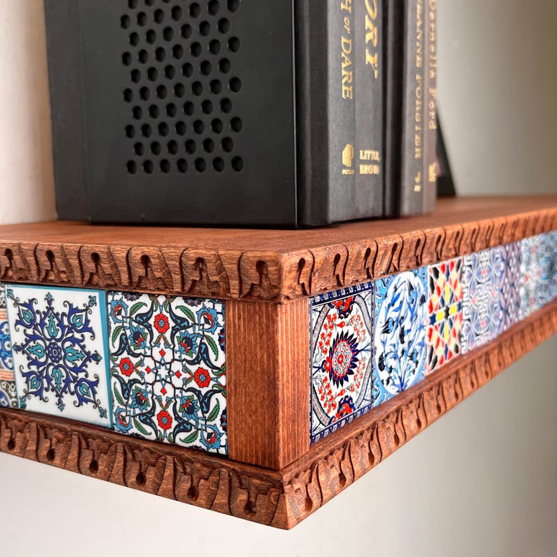 May include: A wooden floating shelf featuring a carved edge with repeating arch patterns and a front panel inlaid with square ceramic tiles displaying intricate blue, red, and white floral and geometric patterns, supporting black books.