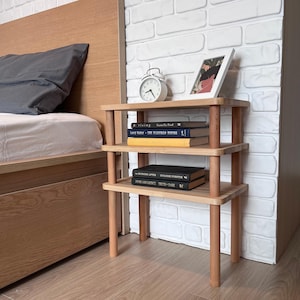 Puede incluir: Una mesita de noche de madera de tres niveles con acabado natural. La mesita tiene forma rectangular con patas y soportes redondeados. Contiene libros, un despertador blanco y una foto enmarcada. La mesita est&aacute; junto a una cama.