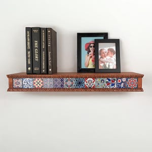 May include: A wooden wall shelf featuring a decorative front edge inlaid with twelve square tiles displaying various colorful floral and geometric patterns. Four black books and two black picture frames are arranged on the shelf surface.