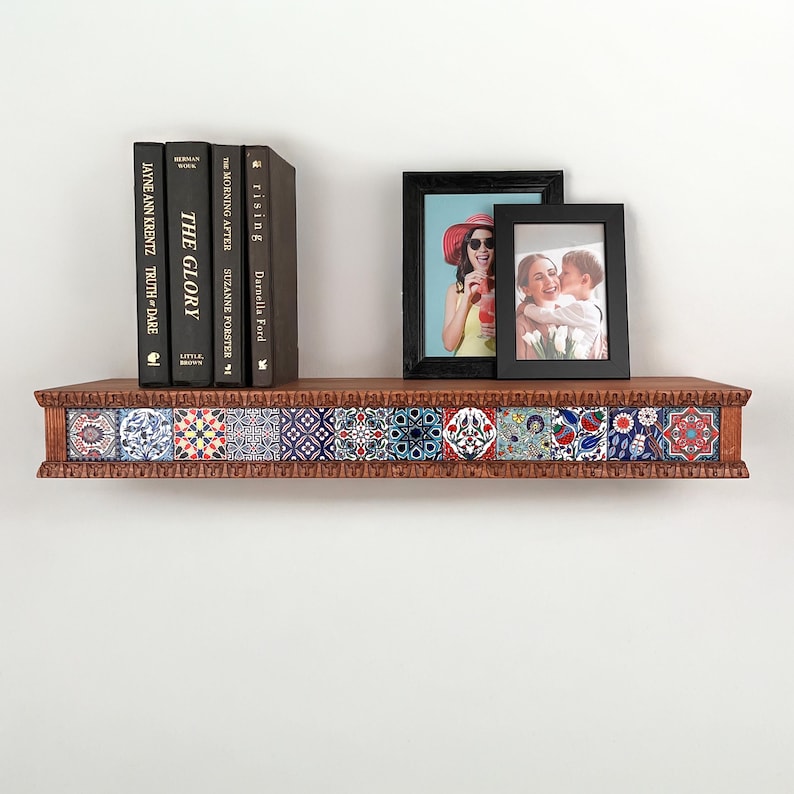 May include: A wooden wall shelf featuring a decorative front edge inlaid with twelve square tiles displaying various colorful floral and geometric patterns. Four black books and two black picture frames are arranged on the shelf surface.