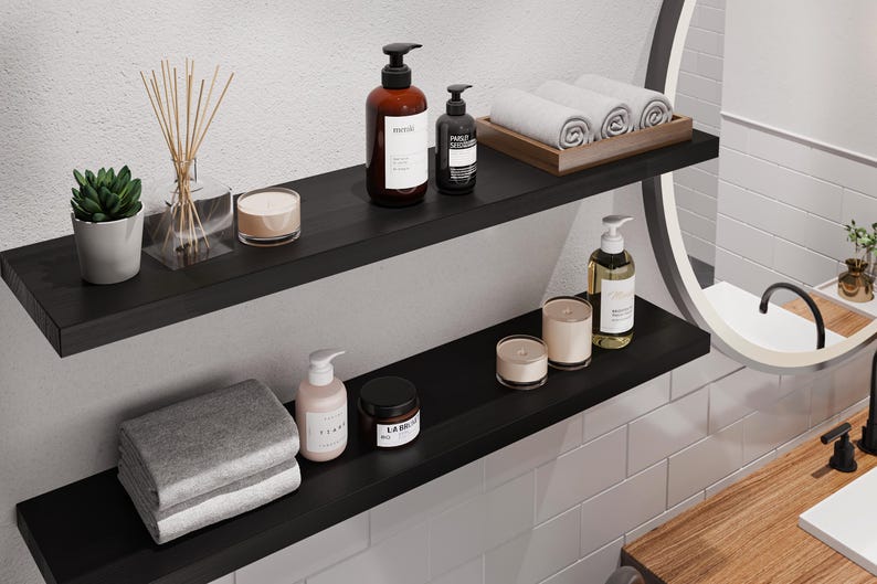 May include: Two black floating shelves with various bathroom items, including two bottles of hand soap, a diffuser, candles, and towels. The shelves are mounted on a white tiled wall.