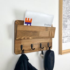 May include: Wooden wall mounted key holder with a mail slot. The key holder has four hooks and a brown finish. The mail slot has a white envelope inside with the word "Envelope" printed on it.