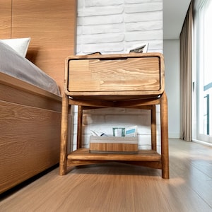 May include: A wooden nightstand with a drawer and a lower shelf. The nightstand is made of light-colored wood and has a rectangular shape. The drawer is closed, and the shelf holds a box. The nightstand is in a bedroom setting.
