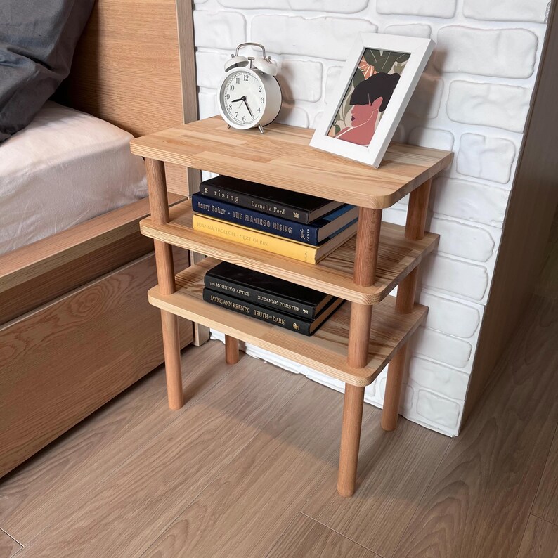 Puede incluir: Una mesita de noche de madera de tres niveles con acabado natural. La mesa tiene una parte superior rectangular y tres estantes sostenidos por patas cil&iacute;ndricas. Libros, un despertador blanco y una imagen enmarcada se exhiben en la mesa. La mesa mide aproximadamente 61 cm de alto.