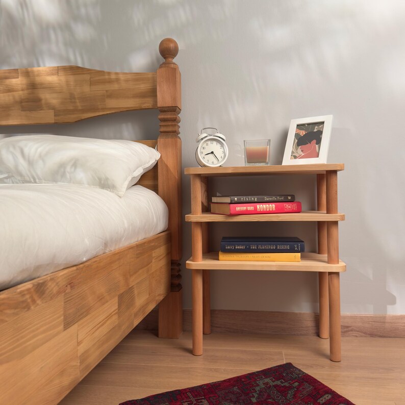 Puede incluir: Una mesita de noche de madera clara con tres estantes, que contiene libros, un peque&ntilde;o despertador blanco, una vela y una foto enmarcada. La mesita de noche est&aacute; junto a una estructura de cama de madera.