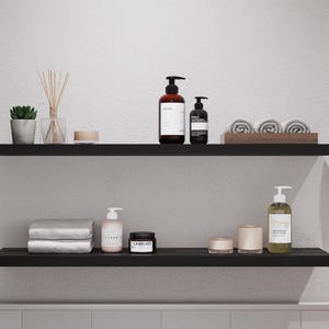 May include: Two black shelves with various bathroom products, including bottles of lotion, a jar of cream, and towels. The shelves are mounted on a white wall.