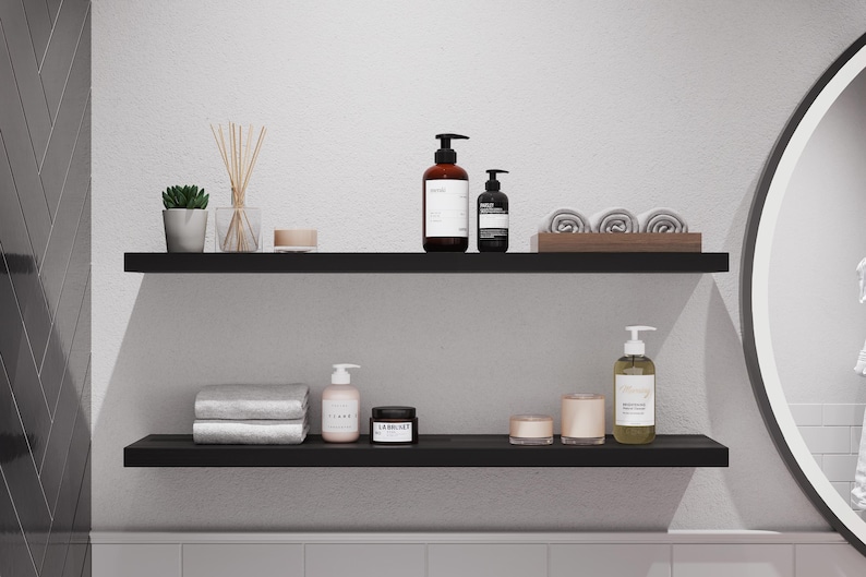 May include: Two black shelves with various bathroom products, including bottles of lotion, a jar of cream, and towels. The shelves are mounted on a white wall.