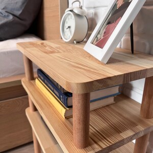 Puede incluir: Una mesita de noche de madera clara con dos estantes. Un despertador blanco y una foto enmarcada est&aacute;n en el estante superior. Los libros est&aacute;n apilados en el estante del medio. La mesita tiene esquinas redondeadas y patas cil&iacute;ndricas.