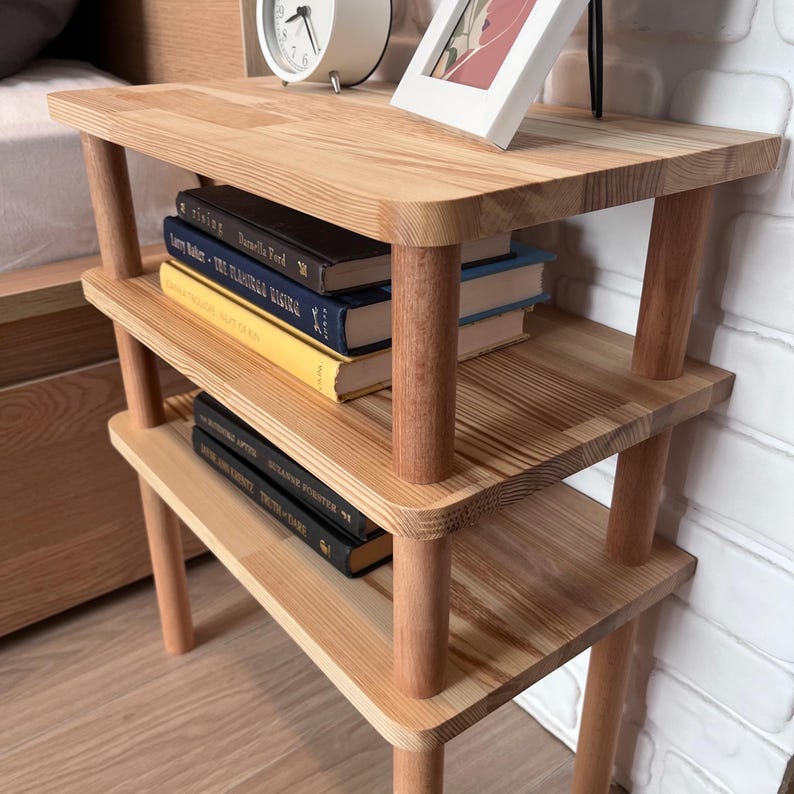 Puede incluir: Una mesita de noche de madera de tres niveles con esquinas redondeadas y patas cil&iacute;ndricas. El acabado de madera natural complementa los libros y objetos decorativos de los estantes. La mesa mide unos 61 cm de alto.