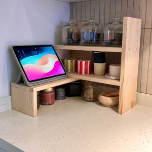 May include: A light-coloured wooden tiered shelf unit with various kitchen items. An iPad displays 09:29. Glass jars with lids, small containers, and a mortar and pestle are visible. The unit sits on a countertop.