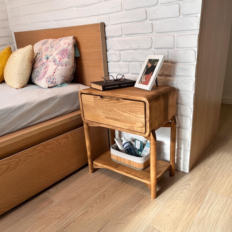 May include: A wooden nightstand with a drawer and a lower shelf. The nightstand is made of light-colored wood and has rounded edges. A book, glasses, and a framed photo sit on top. A basket with toiletries is on the shelf below. The nightstand is next to a bed.
