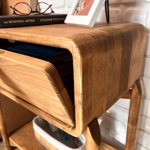 May include: A wooden nightstand with a drawer slightly open, revealing a blue book. The nightstand has a warm, natural wood finish and rounded edges. A book and framed picture sit on top. The nightstand is supported by four legs.