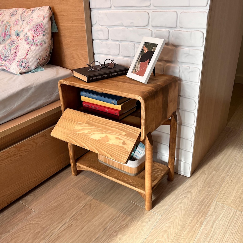 May include: A wooden nightstand with a curved design, featuring a drawer and a lower shelf. The nightstand holds books, a framed picture, and eyeglasses. The wood has a natural finish, and the nightstand is placed next to a bed.