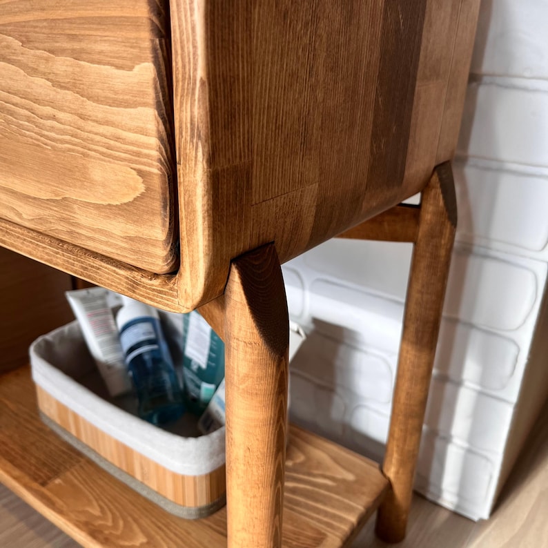 May include: Close-up of a wooden nightstand with a drawer and angled legs. The nightstand is made of light brown wood with visible grain. A small shelf holds a basket with toiletries. The design is minimalist and modern.