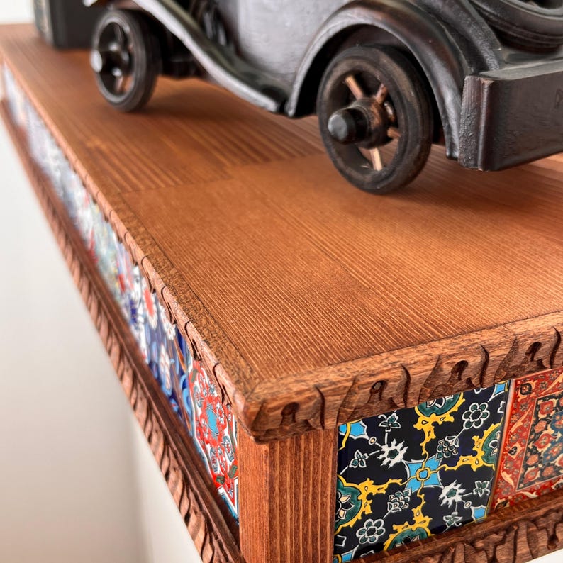 May include: A rectangular wooden shelf with a carved edge and decorative tiles featuring intricate floral and geometric patterns in blue, yellow, red, and black. A black vintage car model sits on the top surface of the wooden shelf.
