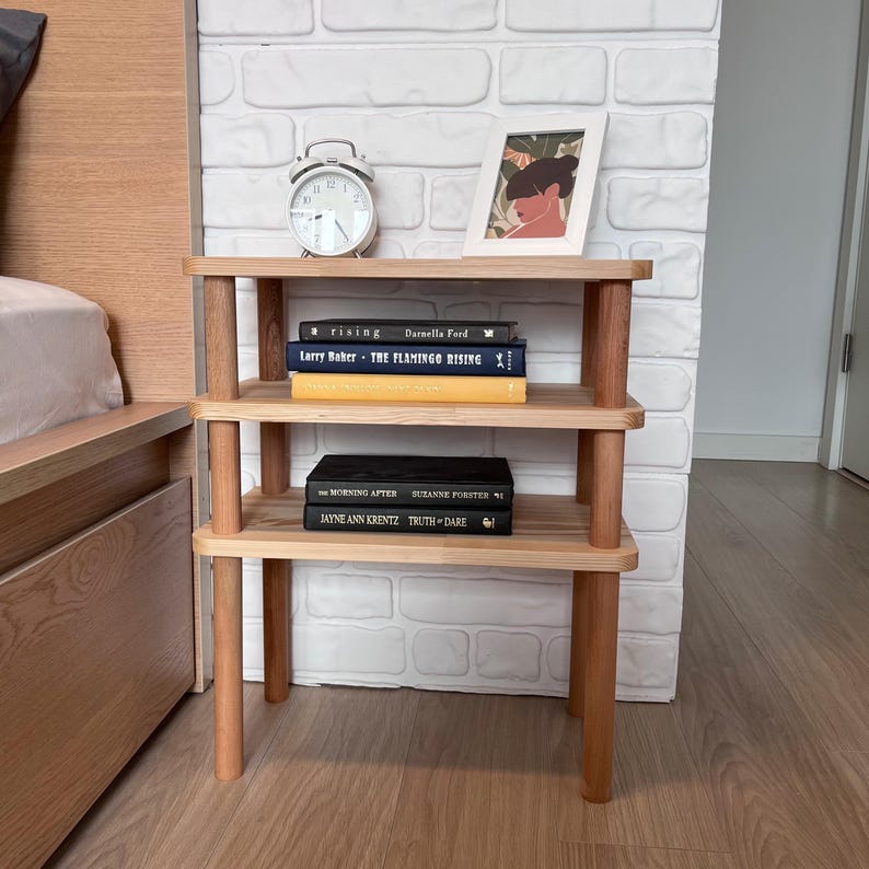 Puede incluir: Una mesita de noche de madera de tres niveles con un acabado natural. La mesita contiene libros, una imagen enmarcada y un despertador blanco. Los libros incluyen t&iacute;tulos como "rising" y "THE FLAMINGO RISING". La mesita es de madera clara.