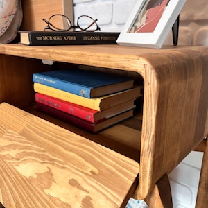 May include: A wooden nightstand with a curved design, featuring a shelf with a stack of books in blue, yellow, and red. The top surface holds a book, glasses, and a framed picture. The nightstand has a warm, natural wood finish.