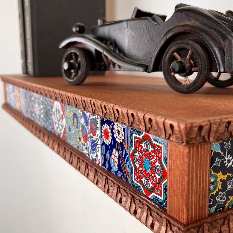 May include: A wooden wall shelf featuring a carved edge and a front panel decorated with a series of square ceramic tiles. The tiles display intricate floral and geometric patterns in vibrant shades of red, blue, white, and yellow. A dark toy car sits on top.