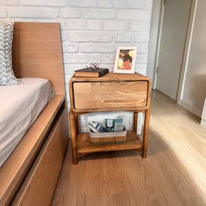 May include: A wooden nightstand with a drawer and a lower shelf. The nightstand is made of light-colored wood and has a rectangular shape with rounded edges. A book, glasses, and a framed picture sit on top. A bed is visible on the left.