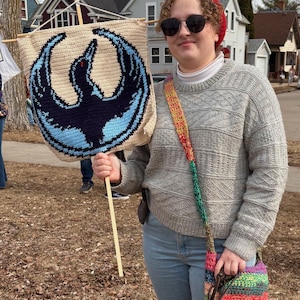 Puede incluir: Una pancarta de ganchillo beige con un emblema de ave azul oscuro y azul claro. La pancarta está sujeta por un palillo de madera. La persona lleva un suéter gris, vaqueros azules y un gorro rojo.