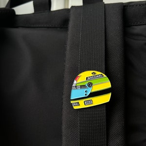 May include: A yellow, black and blue enamel pin of a racing helmet with the words "Nacional" and "Boss" on it.