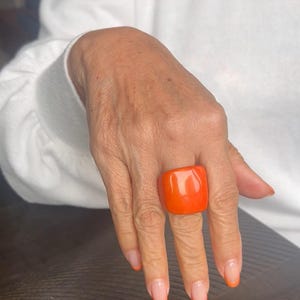 Puede incluir: Un gran anillo cuadrado naranja es el punto focal de esta imagen. El anillo se lleva en una mano con uñas manicuradas, con puntas naranjas. La persona lleva un suéter blanco.