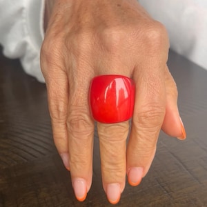 May include: A bold, red square ring is displayed on a finger. The hand is visible, with nails featuring a French manicure with orange tips. The ring is the main focus, with a glossy finish.