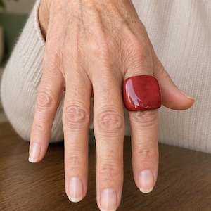 May include: A ring featuring a large, smooth, square-shaped red stone with rounded edges. The stone has a glossy, reflective finish and is set on a simple band, worn on the ring finger. The design is bold, solid, and monochromatic in colour.