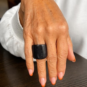 May include: A large, square black ring is worn on a hand with orange-tipped nails. The ring is the focal point, set against a white long-sleeved top and a dark wooden surface.