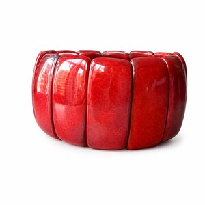 Può includere: Un bracciale elastico composto da molteplici segmenti rettangolari rosso lucido con una finitura liscia e levigata. I singoli pezzi sono collegati tra loro per formare una fascia continua e flessibile di forma arrotondata.