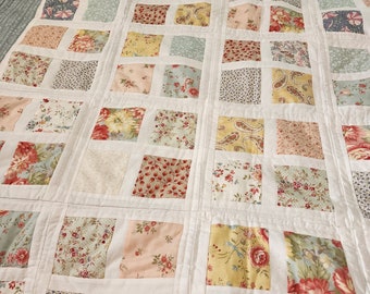 May include: A patchwork quilt with a grid pattern of fabric squares in various floral and paisley prints. The squares are in shades of red, pink, yellow, and blue, set against a white background. The quilt has a light blue border.
