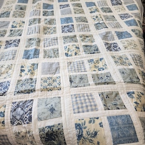 May include: A blue and white patchwork quilt featuring a variety of floral and geometric patterns. The quilt is made up of squares and rectangles, and the fabric is a mix of cotton and linen.