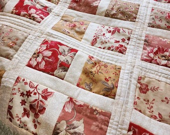 May include: A patchwork quilt with a floral pattern in shades of red, cream, and tan. The quilt is made of rectangular blocks, each featuring a different floral design. The blocks are separated by white fabric and stitched together with a quilted pattern.