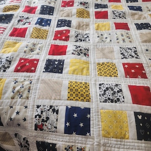 May include: A patchwork quilt featuring a Mickey Mouse theme. The quilt is made up of squares of different fabrics, including red, white, blue, yellow, and black. The squares are arranged in a random pattern, and some of the squares feature Mickey Mouse characters.