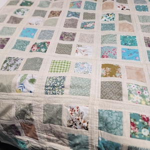 Puede incluir: Una colcha beige con un diseño de patchwork que presenta una variedad de patrones florales y geométricos en tonos de verde, azul y marrón. La colcha está compuesta por cuadrados, cada uno con una tela diferente.