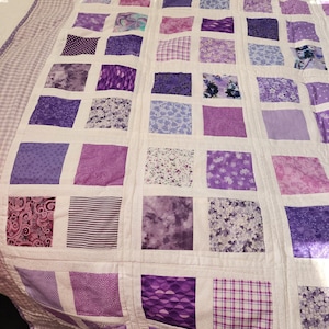 May include: A purple and white patchwork quilt featuring a variety of different patterns. The quilt is made up of squares of different fabrics, all in shades of purple.
