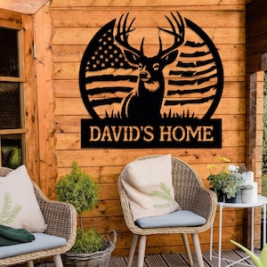 Peut inclure: Décoration murale en métal noir représentant une silhouette de cerf avec des bois, debout devant un motif de drapeau américain. Le texte "DAVID'S HOME" est affiché sous le cerf.
