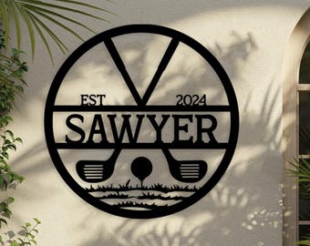 May include: Black metal golf-themed wall art. The design features a circular frame with the name "SAWYER" and the year "2024". Two golf clubs flank a golf ball on a tee, above a grassy area. The word "EST" is above the name.