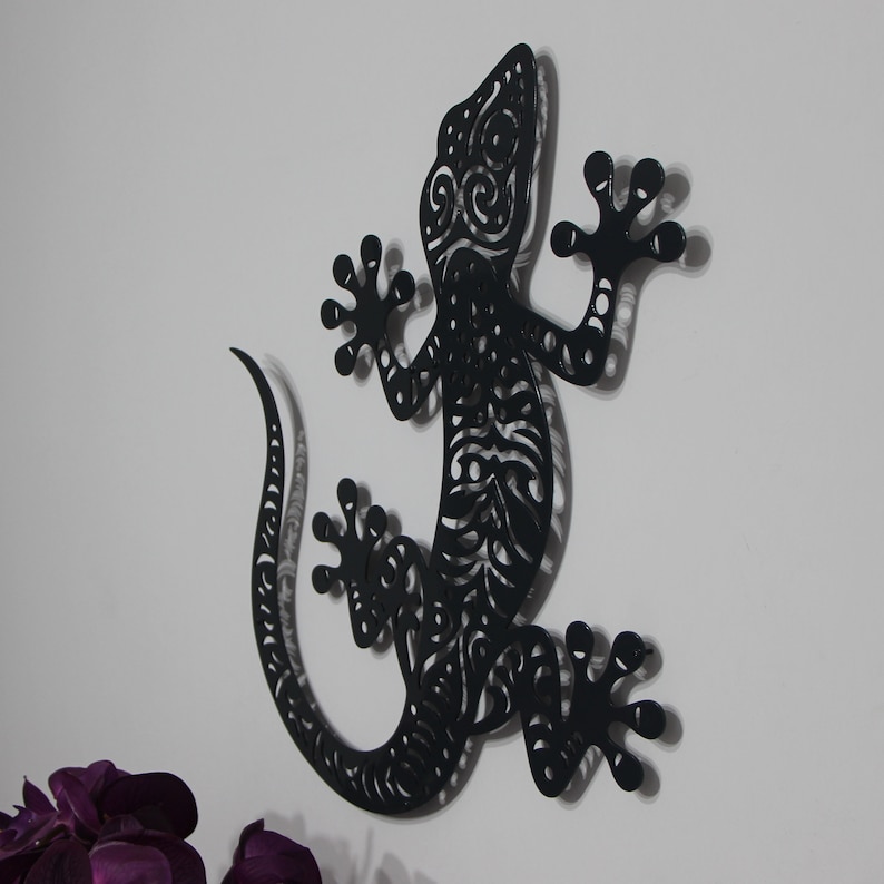 May include: Black metal gecko wall decor with intricate cut-out details. The gecko is posed with its tail curled and its front legs raised.