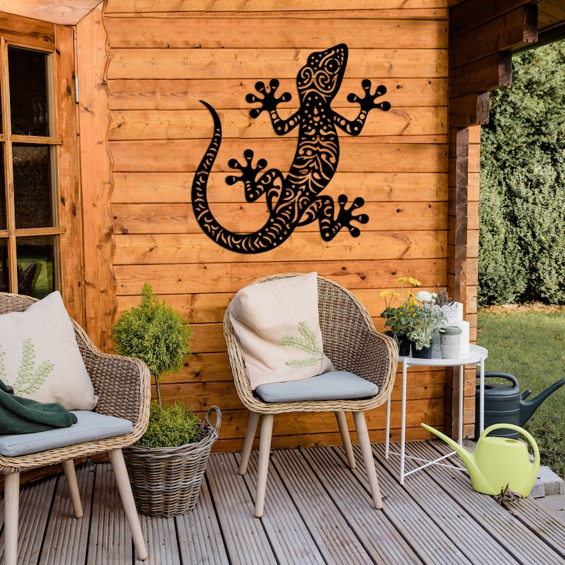 May include: Black metal gecko wall art with intricate details, hanging on a wooden wall with two wicker chairs and potted plants in front of it.