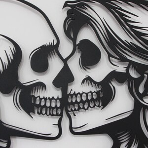 May include: A black laser-cut wall art piece featuring two skulls facing each other in a kissing position. The skull on the right is adorned with detailed hair and a rose. The design is composed of solid black lines against a plain white background.